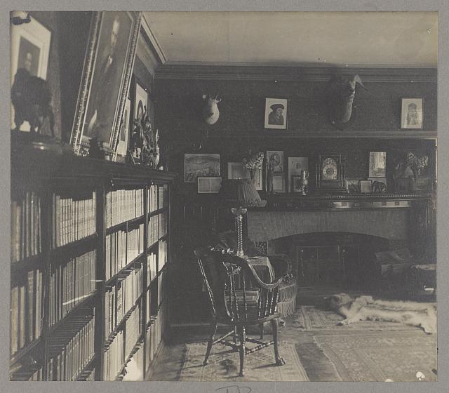  interior of Roosevelt's library at Sagamore Hill with a bookcase, hearth and mantle, several photographs, sculpture, mounted antelope heads, a badger skin rug and other carpeting, and chairs. 1905
